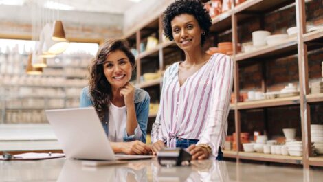 Two women smile at the camera with a laptop and a card reader on the table and shelves in the background.