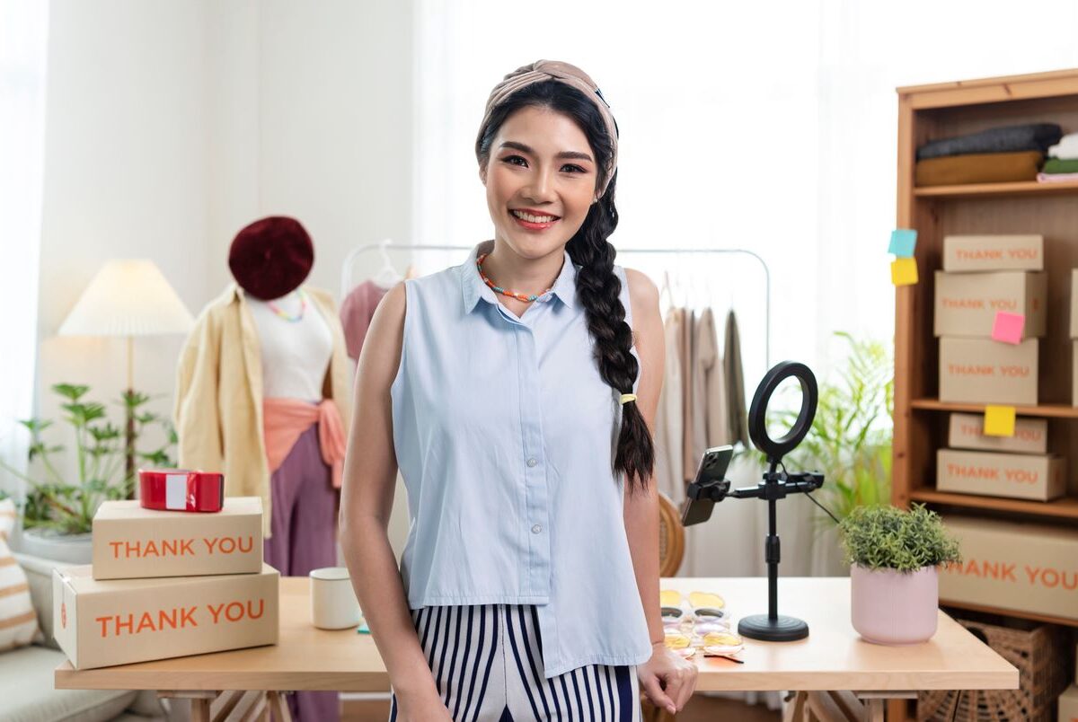 A young small business owner smiles in front of a desk with shipping boxes behind.